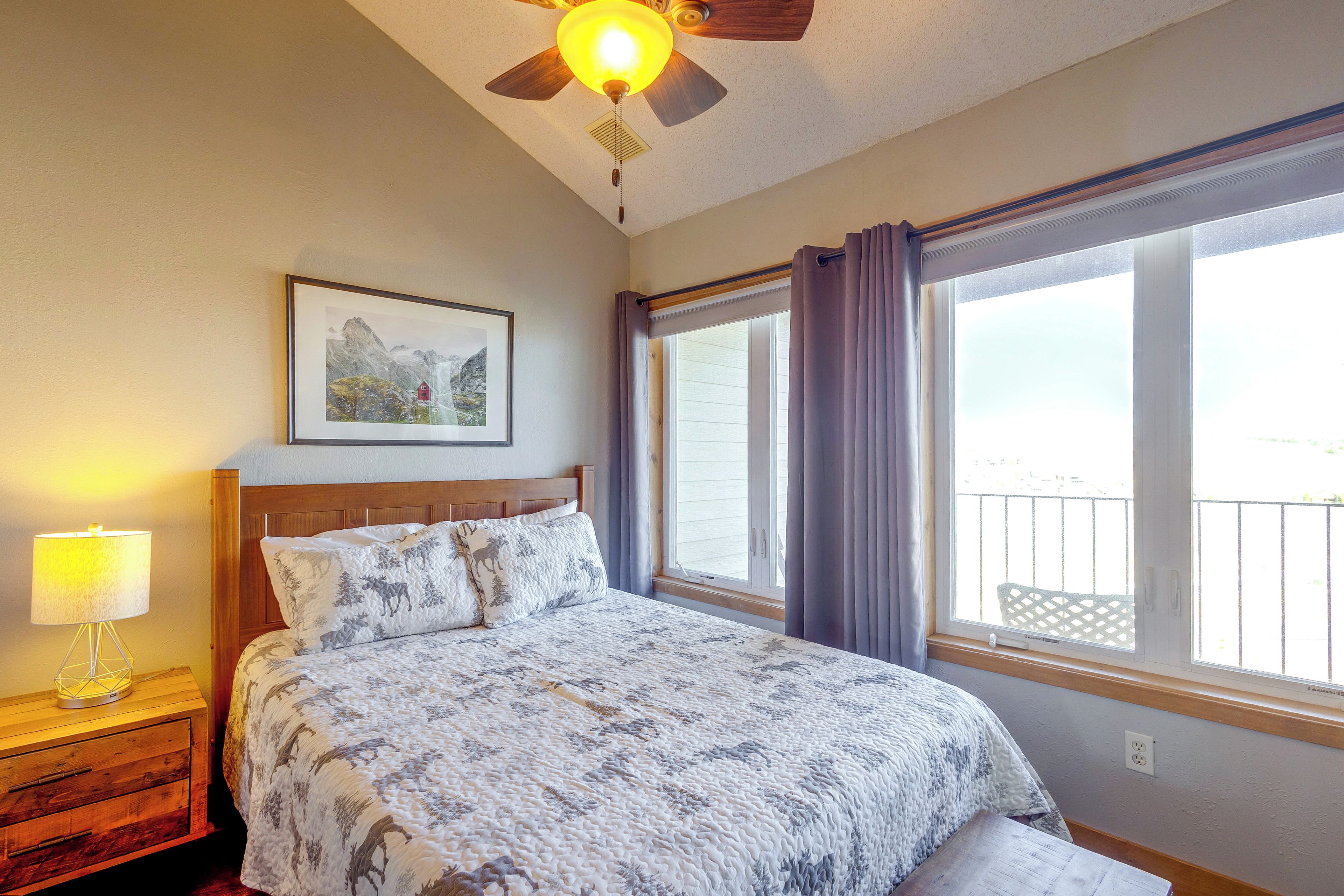 A bedroom with a bed, a nightstand, a lamp, a ceiling fan, a window with curtains, and a framed picture on the wall.