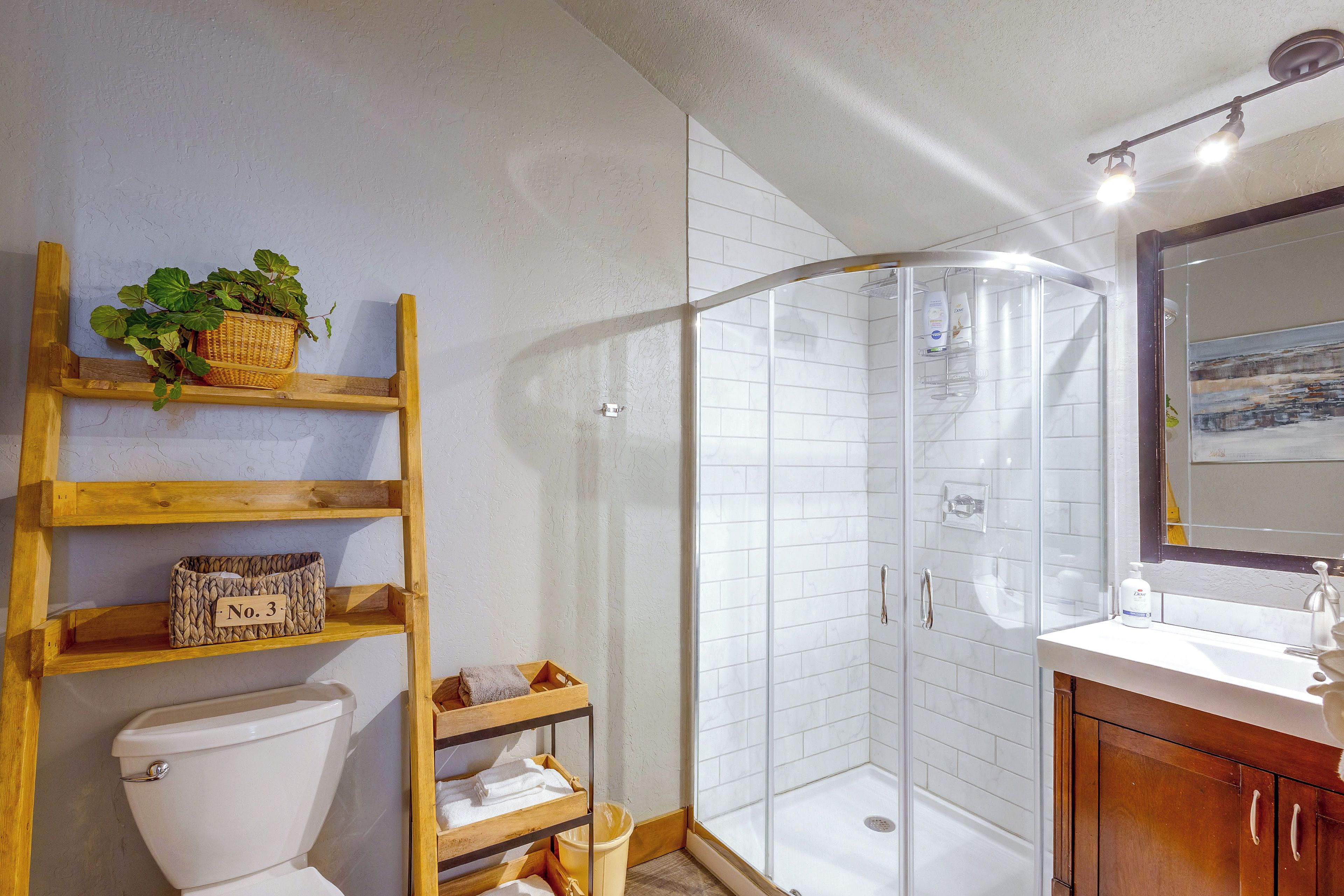 Un baño con inodoro, un estante de madera con una cesta y una caja etiquetada 'No. 3', cerramiento de ducha de vidrio y un lavamanos con espejo.