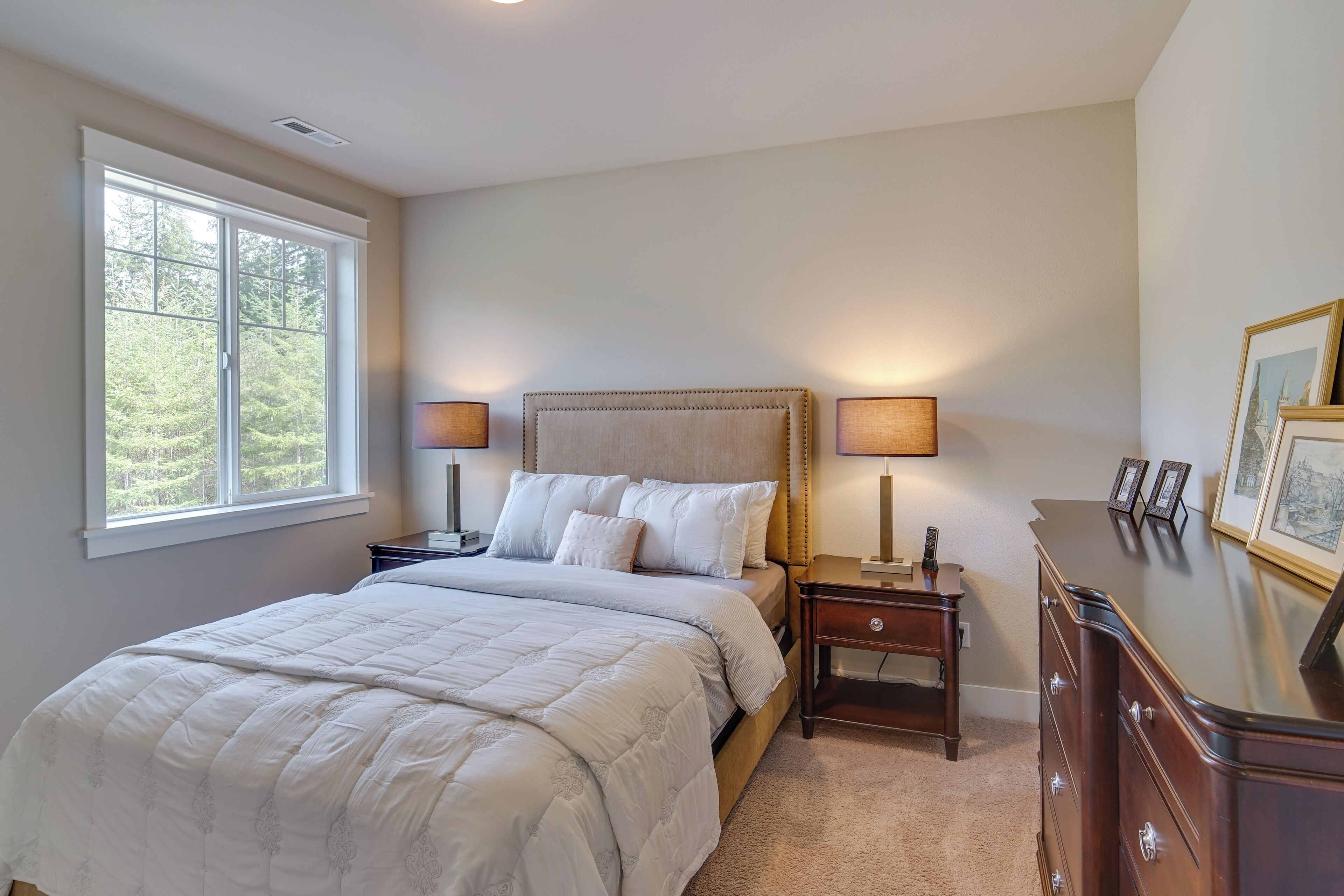 A bedroom with a large bed, two bedside tables with lamps, a dresser, and a window with a view of greenery.
