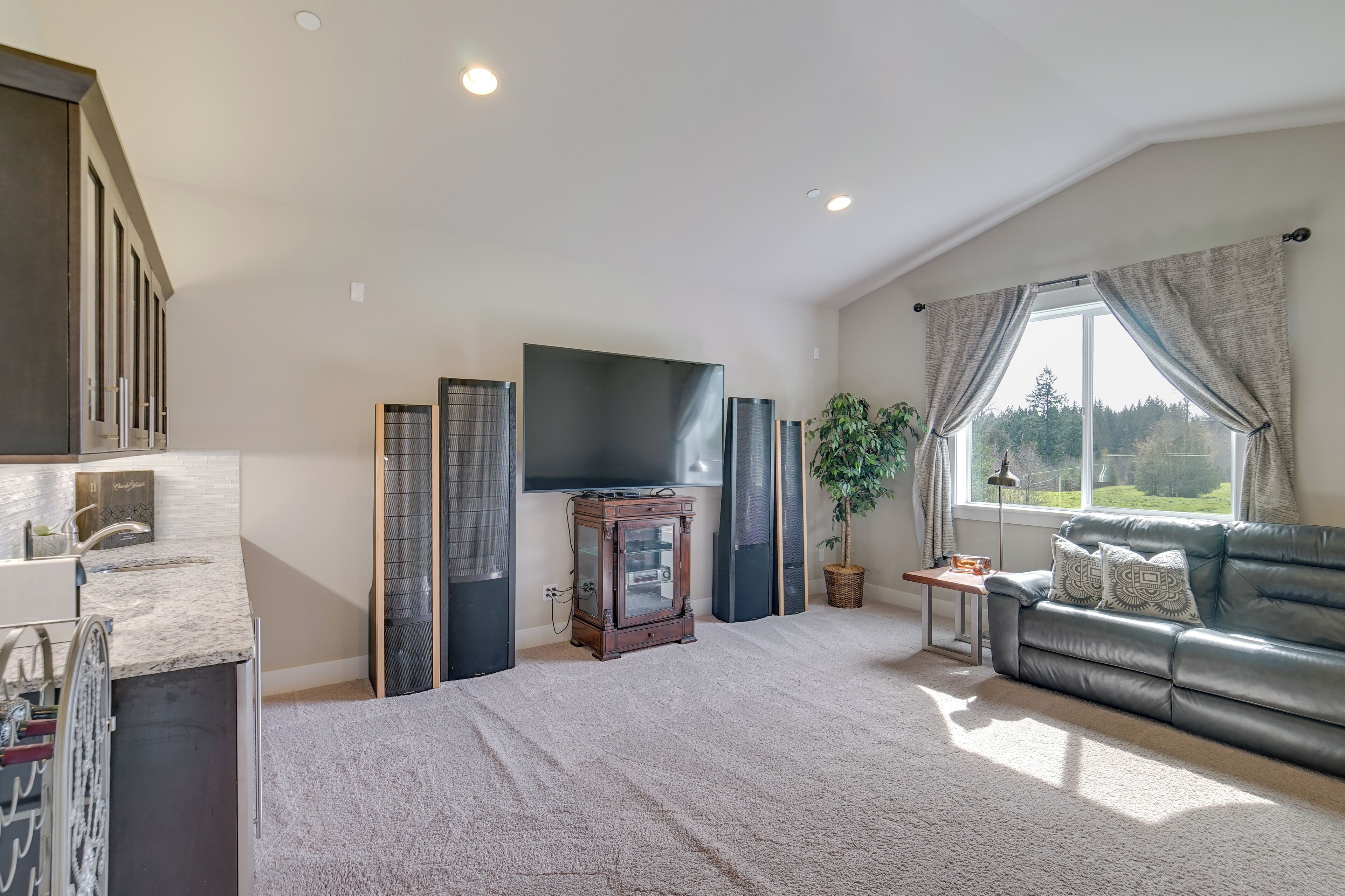 A spacious living room with a large window, a dark leather sofa, a flat-screen TV, and a small wooden cabinet.