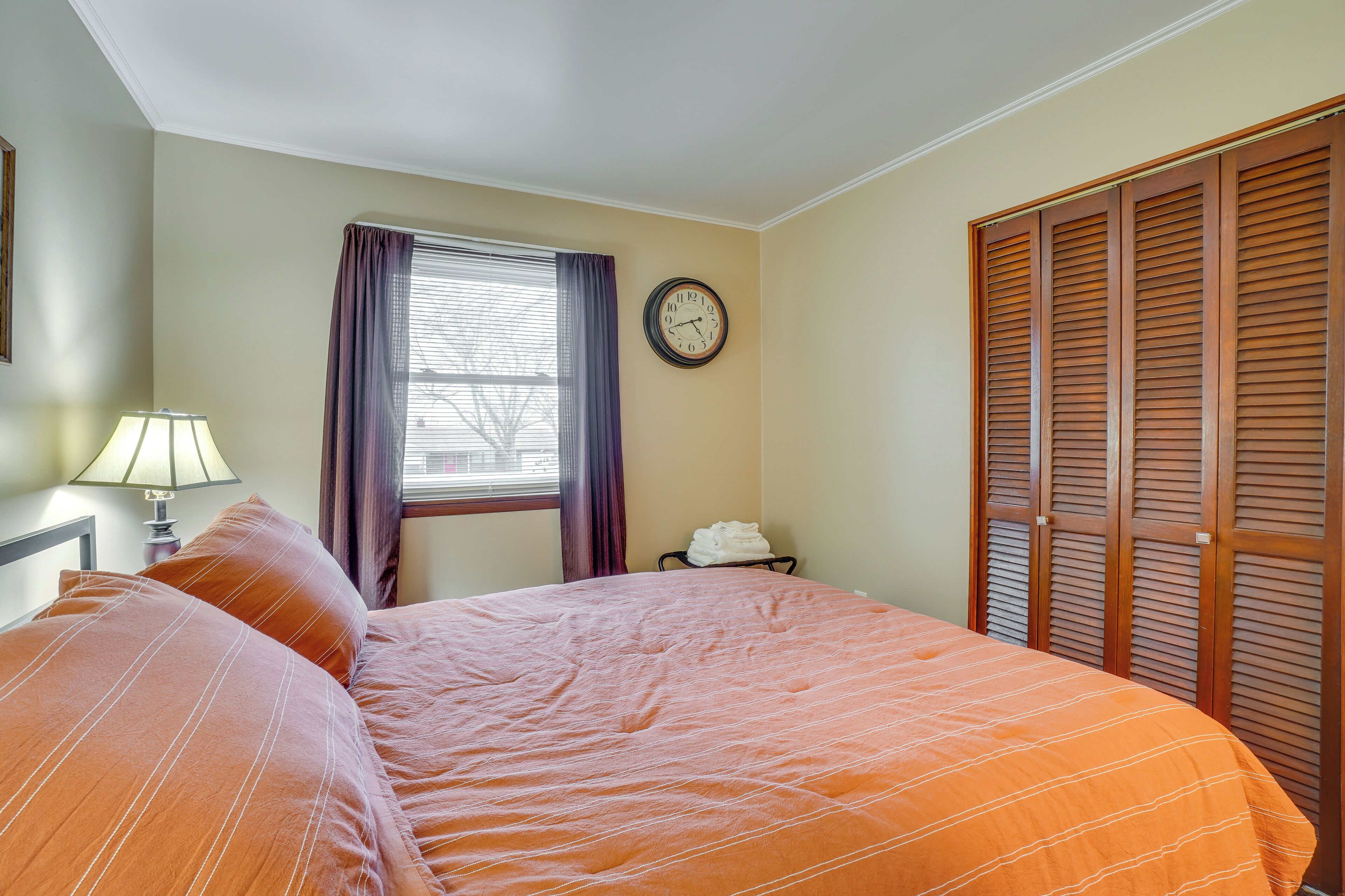 Una cama bien hecha con una colcha naranja, un reloj en la pared y una ventana con cortinas.