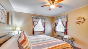A neatly made bed with a striped blanket, two windows with curtains, a ceiling fan, and a wall decoration. - Shell Knob Home w/ Fire Pit, Near Table Rock Lake! (Shell Knob)