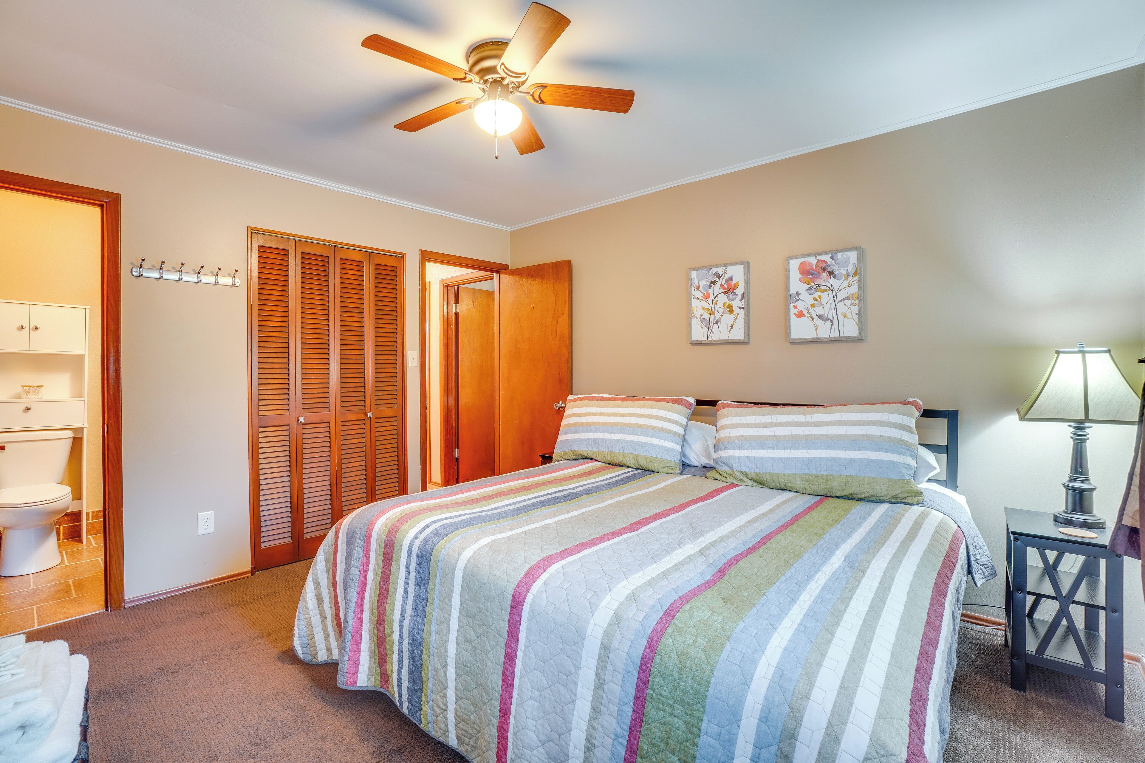 Une chambre à coucher avec un lit, un ventilateur de plafond, une table de chevet, une lampe et une salle de bain visible à travers une porte ouverte.
