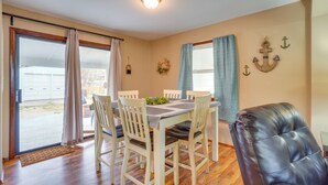 A dining area with a table set for four, surrounded by white chairs, a leather sofa, and a view of the outdoors through large sliding doors. - Shell Knob Home w/ Fire Pit, Near Table Rock Lake! (Shell Knob)