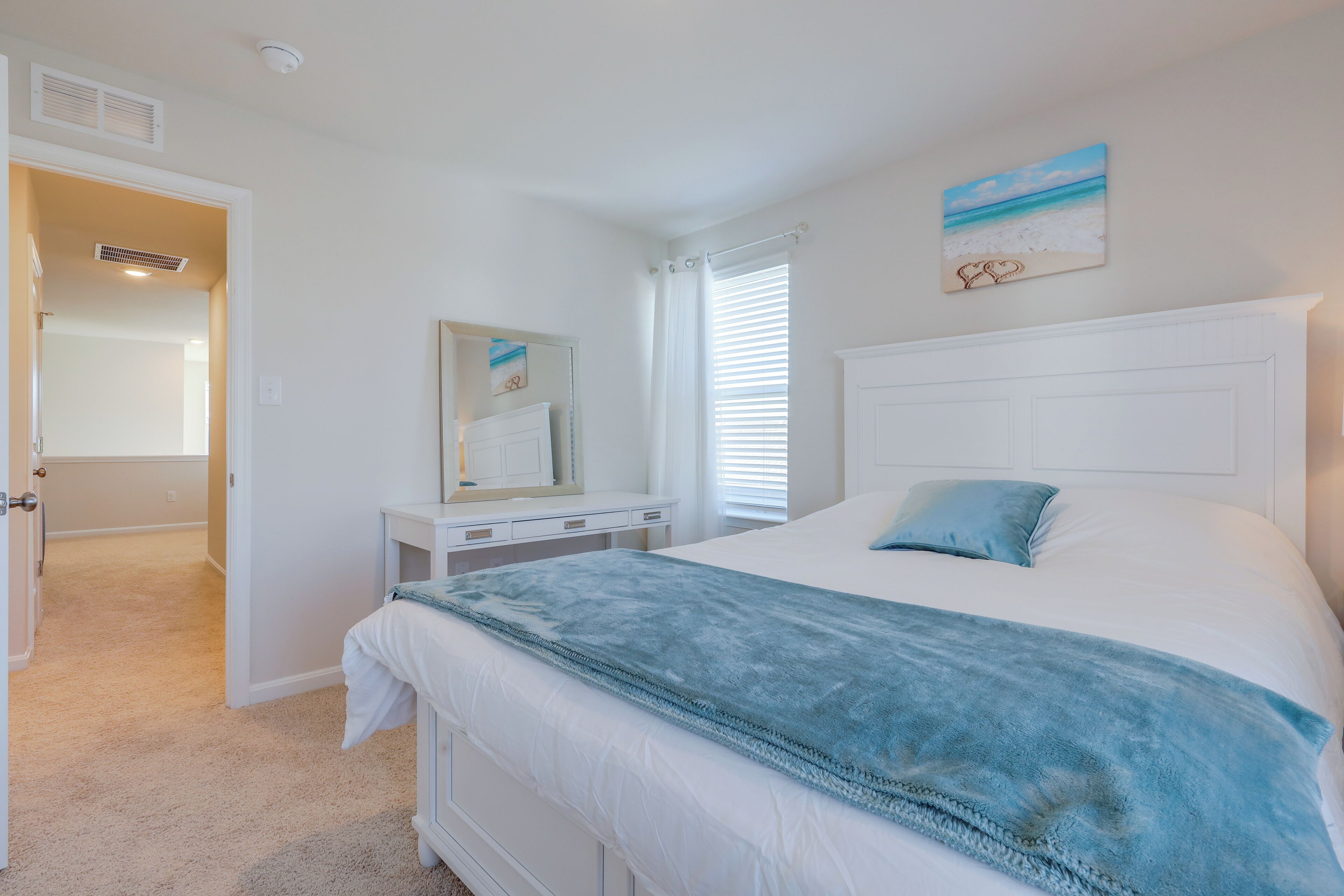 Een slaapkamer met een groot bed, een dressoir, een spiegel en een schilderij van een strandgezicht.