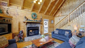 A cozy log cabin interior with a stone fireplace, wooden walls, a blue sofa, and a television. - Smoky Mountains Cabin w/ Hot Tub, Deck & Views! (Sevierville)