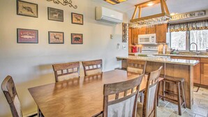 A dining area with a wooden table and chairs, a kitchen with wooden cabinets, and wall-mounted pictures. - Ski Condo w/ Pool, Close to Story Land & Attitash! (Glen)