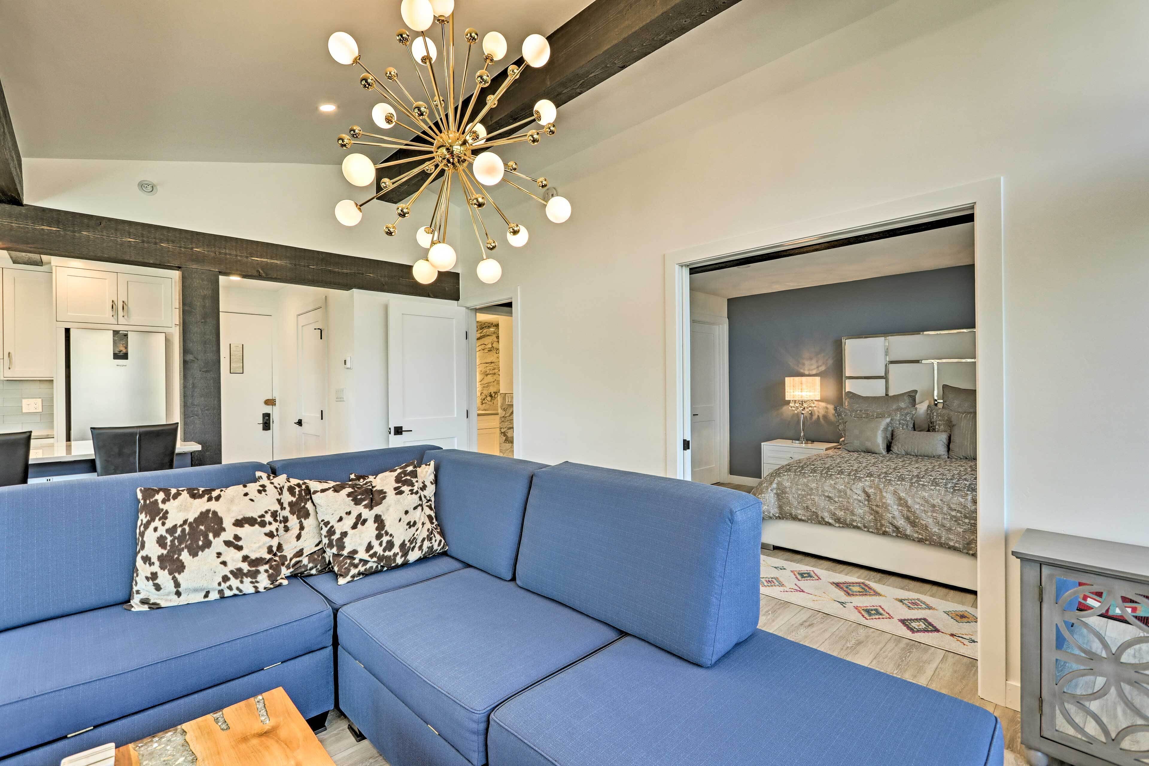 A modern living room with a blue sofa, a unique chandelier, and a bedroom with a bed and nightstand.
