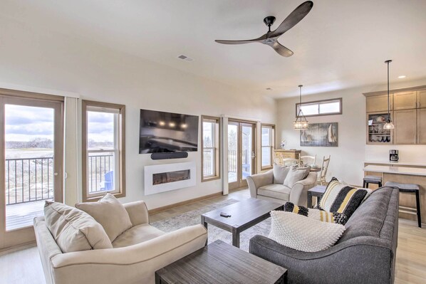 A spacious living room with a fireplace, large windows, a ceiling fan, and a kitchen area with cabinets and pendant lights. - Spacious Recently Built Home < 1 Mi to Lake! (Malone)