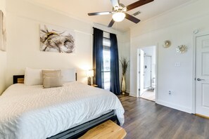 A bedroom with a bed, ceiling fan, artwork, and a view into the bathroom.