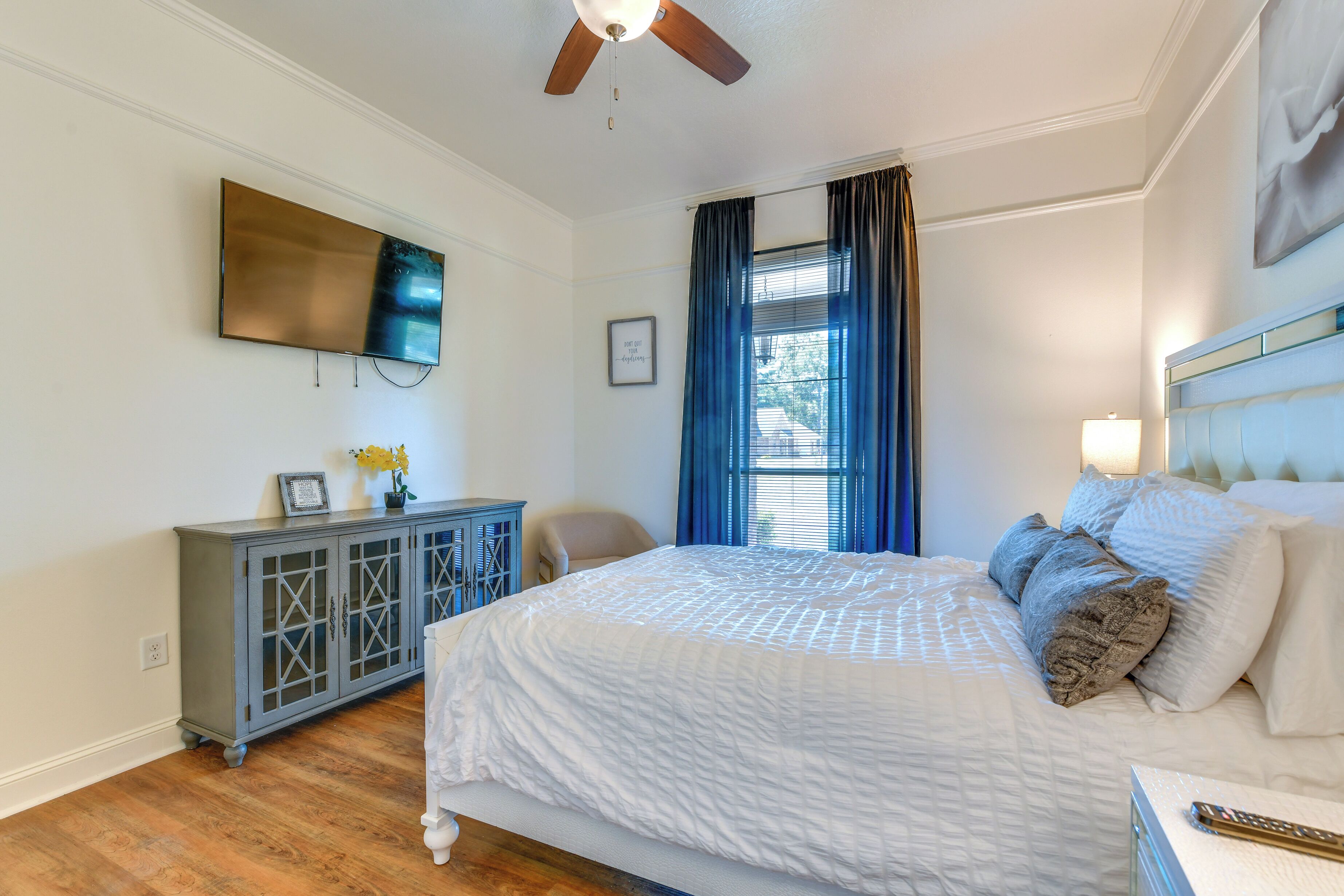 A bedroom with a large bed, a ceiling fan, a TV mounted on the wall, a dresser, and a window with curtains.