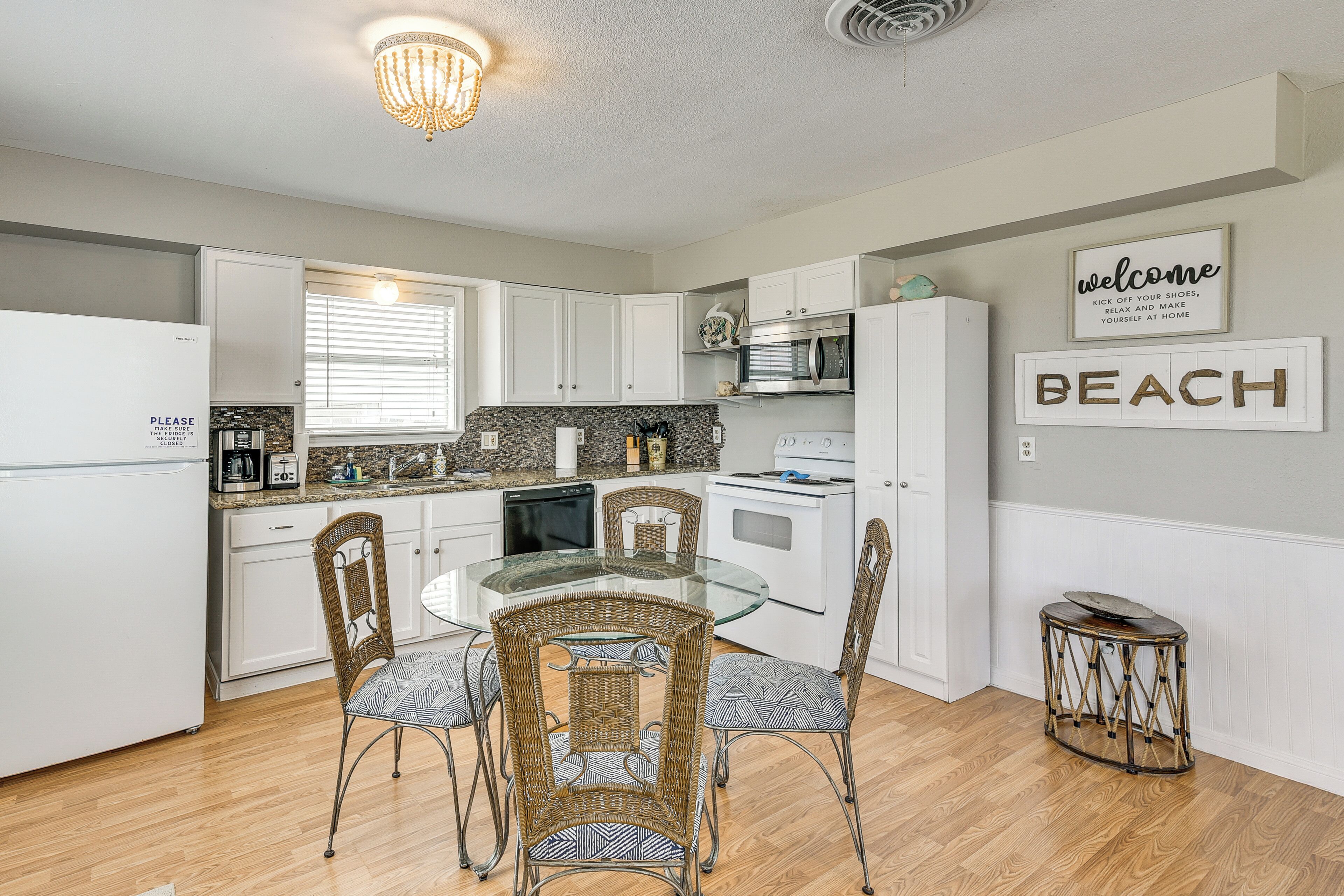 A modern kitchen with white cabinets, a glass dining table, wicker chairs, and a 'BEACH' sign on the wall.