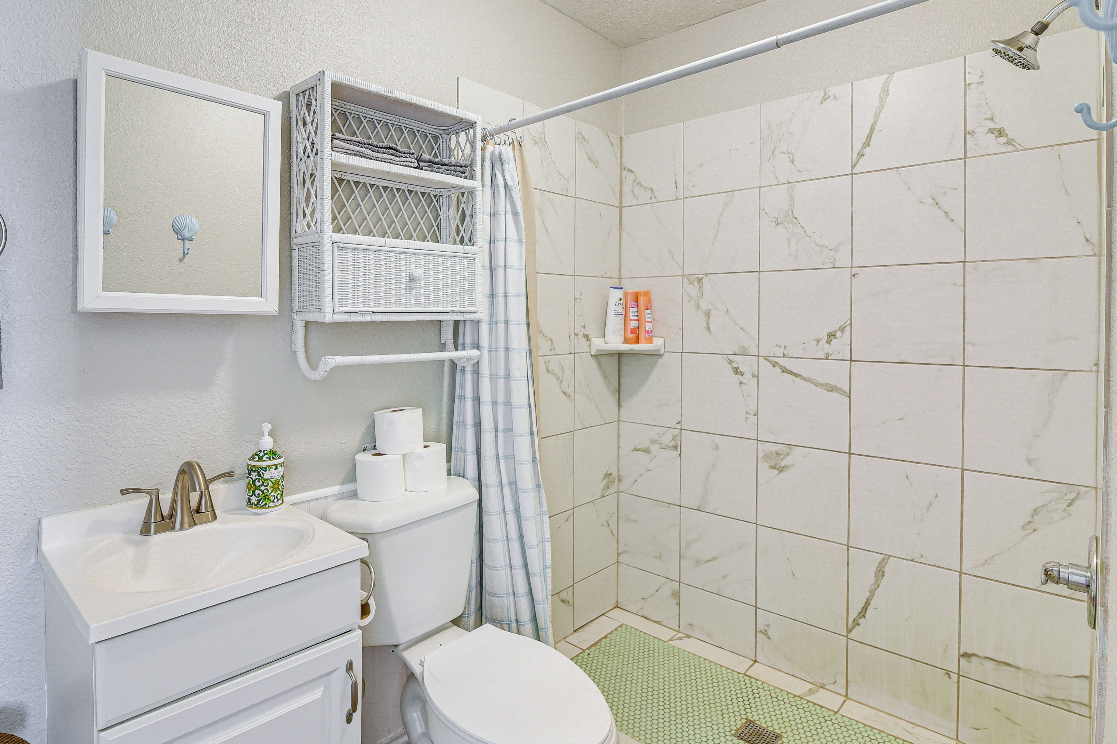 A bathroom with a white sink, toilet, and shower. There is a mirror, a towel rack, and a shelf with toiletries.