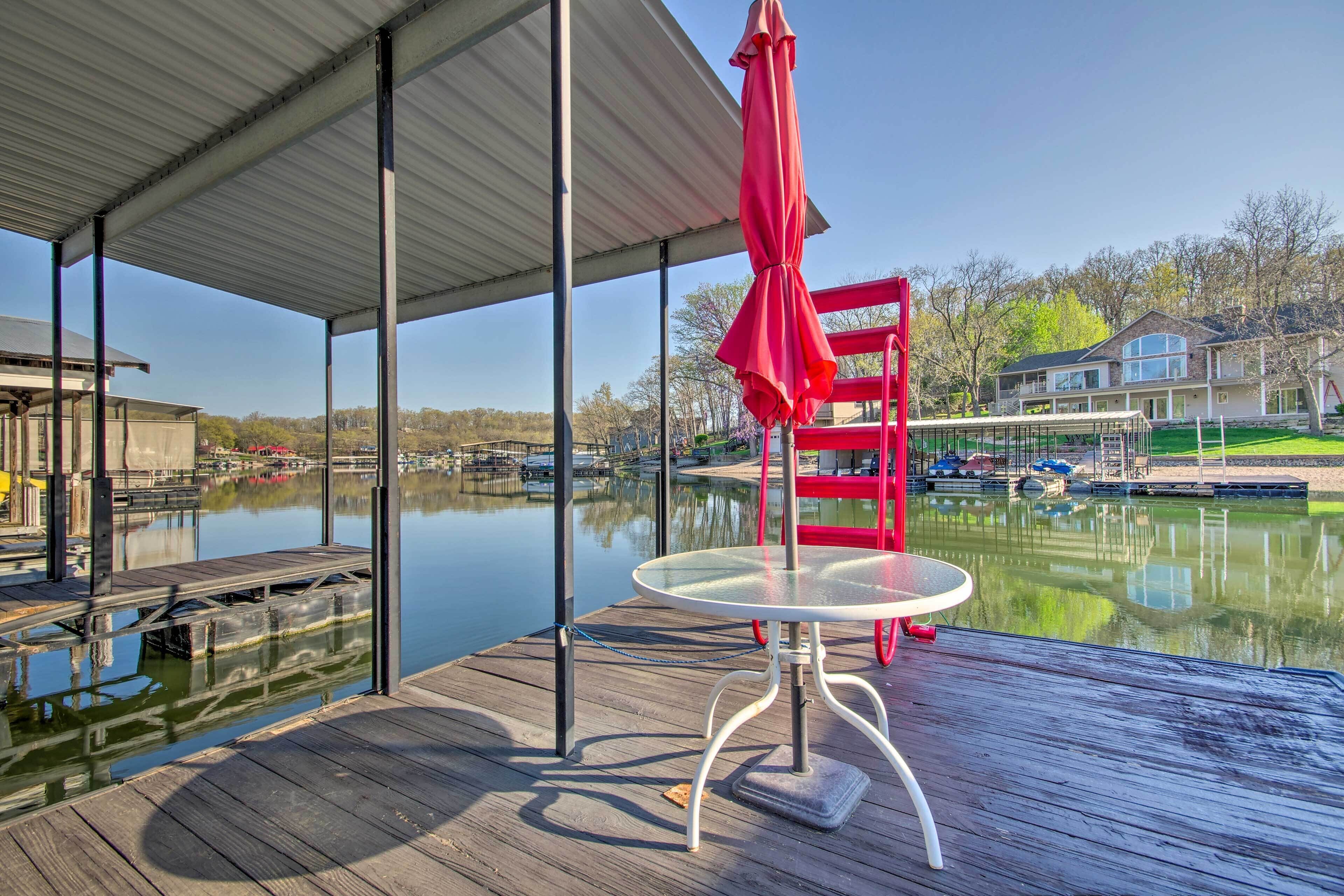 A covered outdoor area with a table and red chair, overlooking a calm body of water and a house in the background.