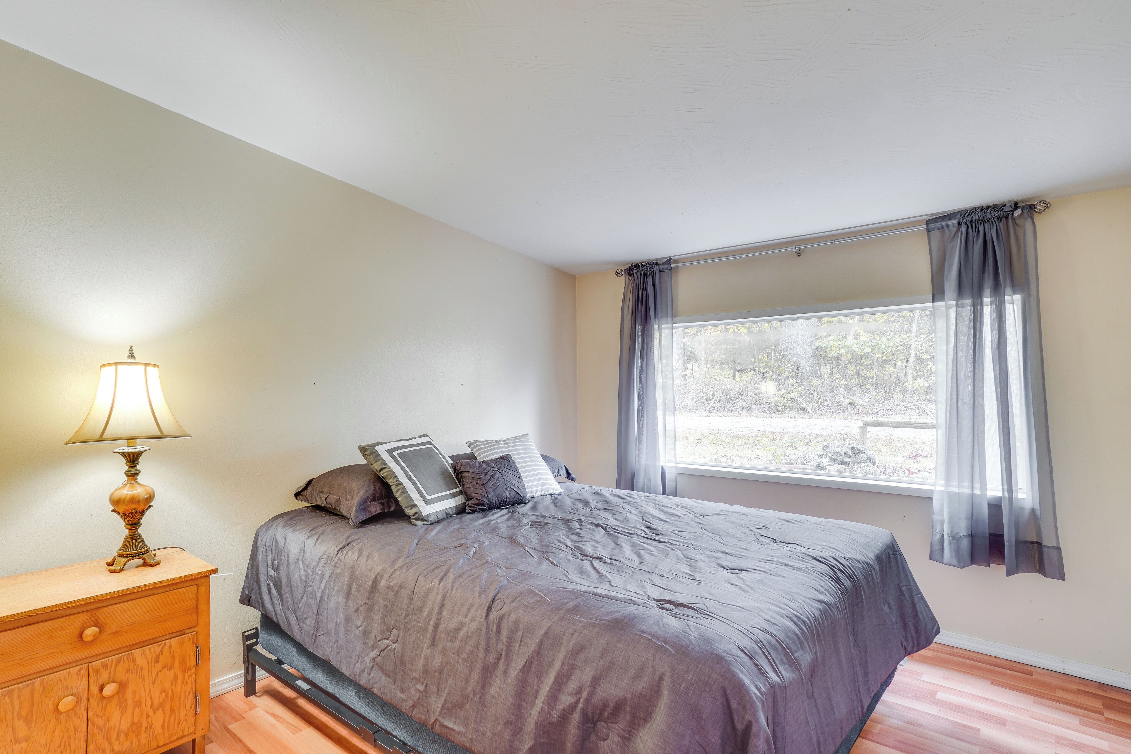 Une chambre à coucher avec un lit, une table de chevet en bois, une lampe, une fenêtre avec des rideaux et une vue sur l’extérieur.