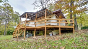 A wooden decked house with a staircase leading to an upper level, surrounded by trees and fallen leaves. - Table Rock Lake Cabin w/ Deck & Bar! (Shell Knob)
