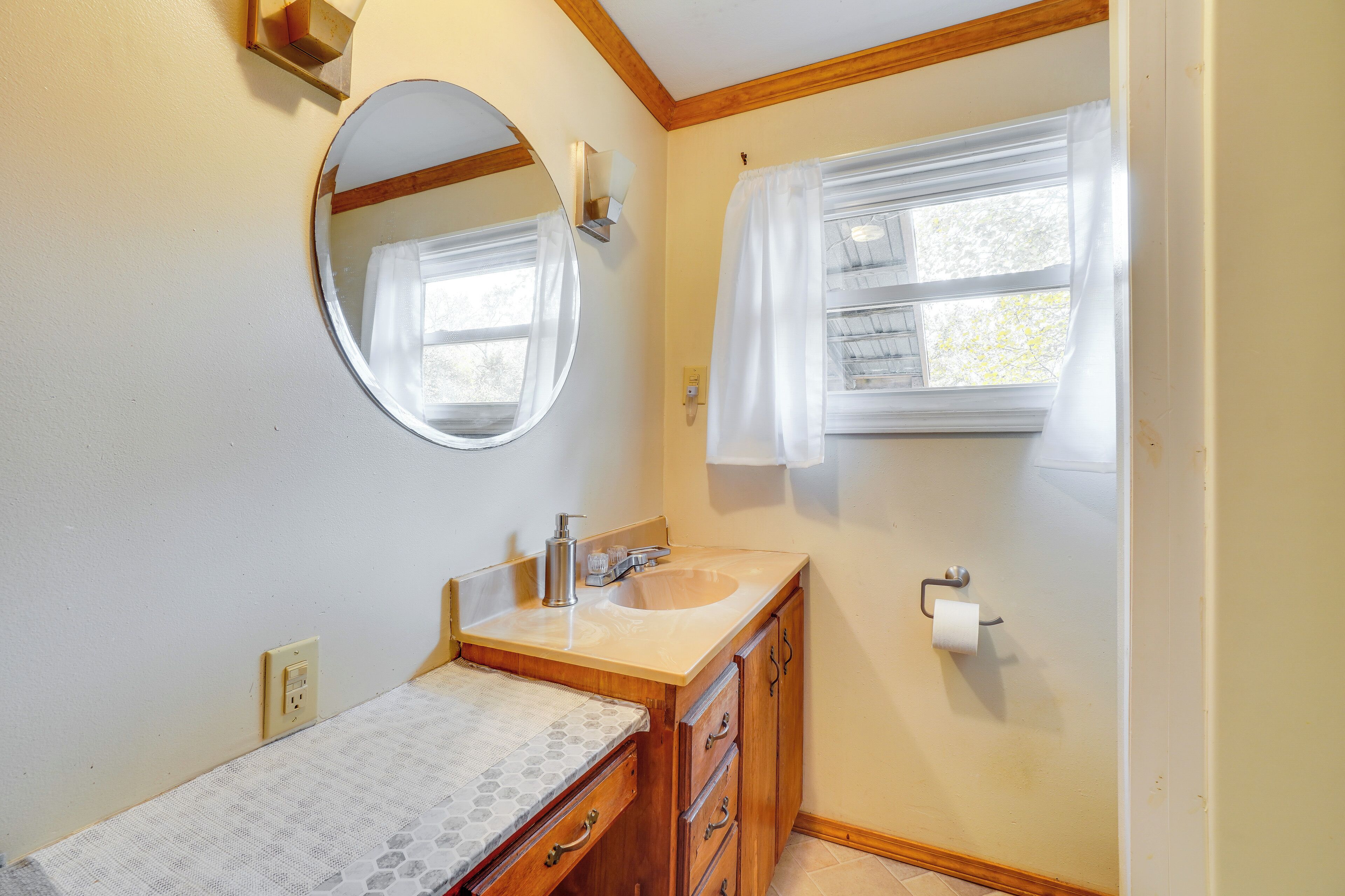 Une salle de bain avec un meuble-lavabo en bois, un miroir rond, un lavabo et une fenêtre avec des rideaux.