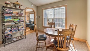 Un comedor con una mesa y sillas de madera, una estantería con juegos de mesa y una ventana con persianas.