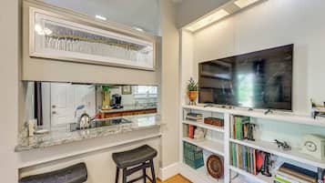 Una cocina con mostrador, lavamanos y dos taburetes. Se observa una estantería con varios libros y objetos decorativos.