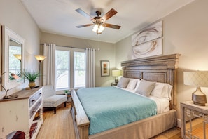 A bedroom with a bed, a desk, a chair, a ceiling fan, and artwork on the wall.