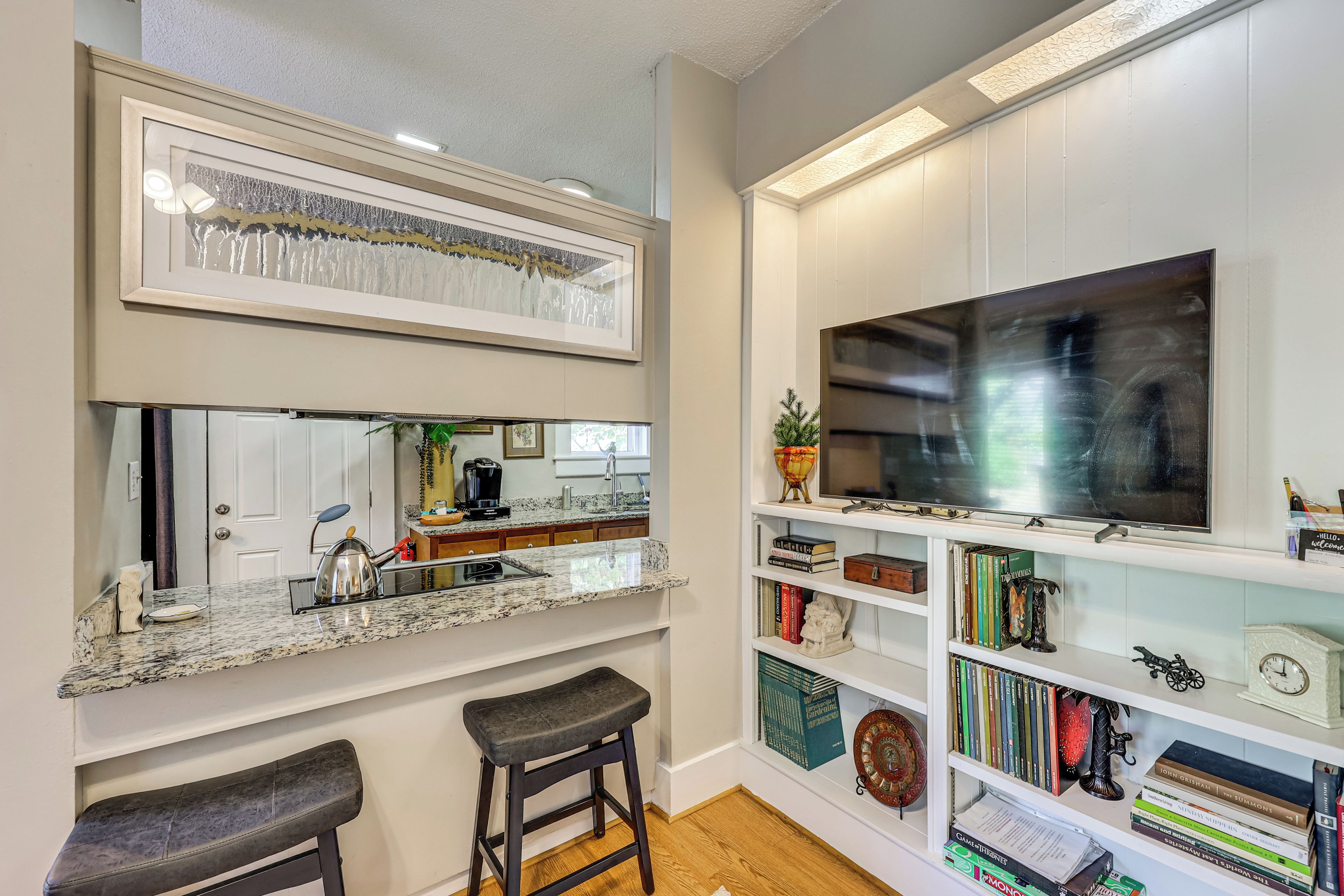Cucina con un piano di lavoro, lavello e due sgabelli. Si nota una libreria con diversi libri e oggetti decorativi.