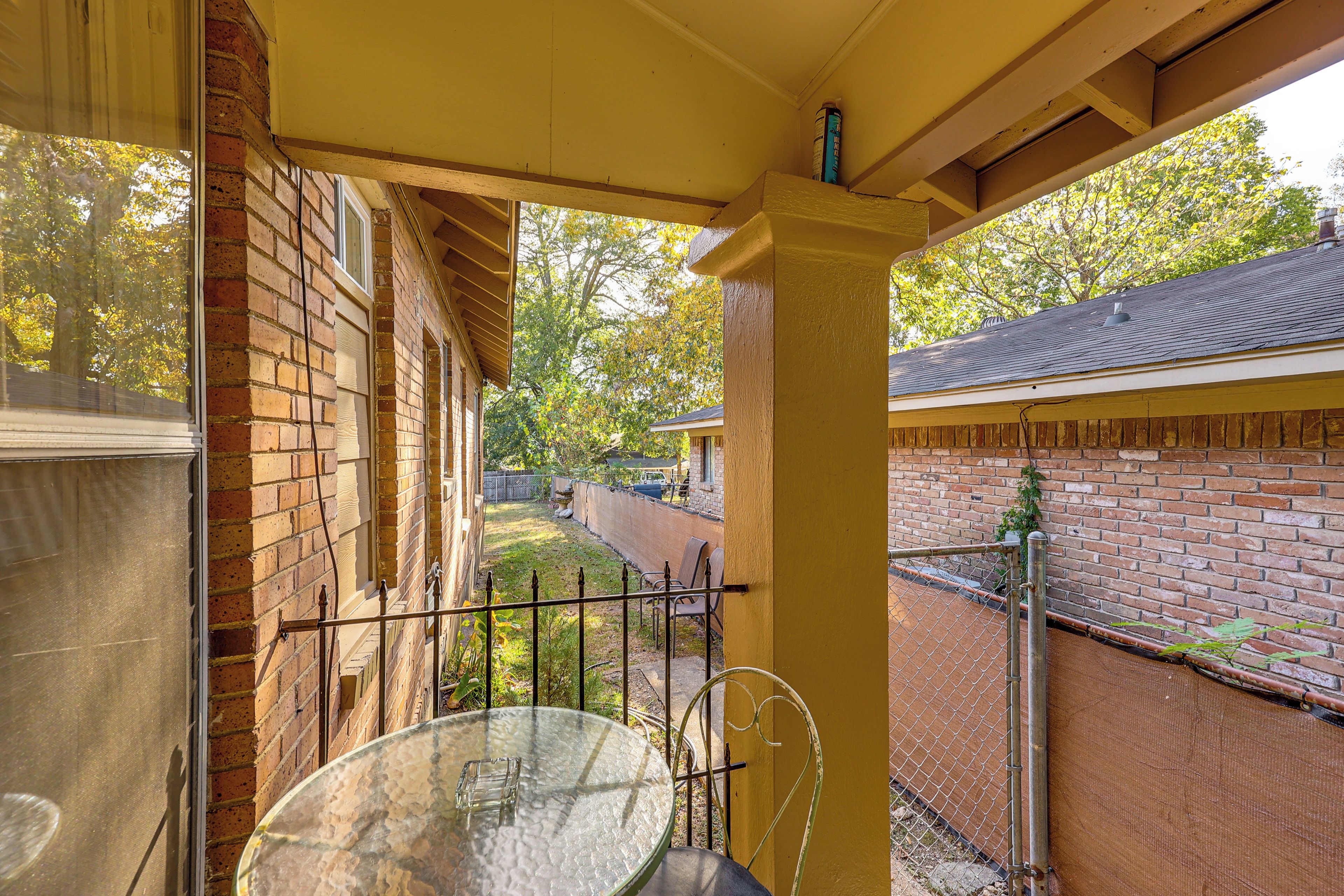 A covered patio area with a glass table and chairs, brick walls, and a view of a backyard with a fence and trees.