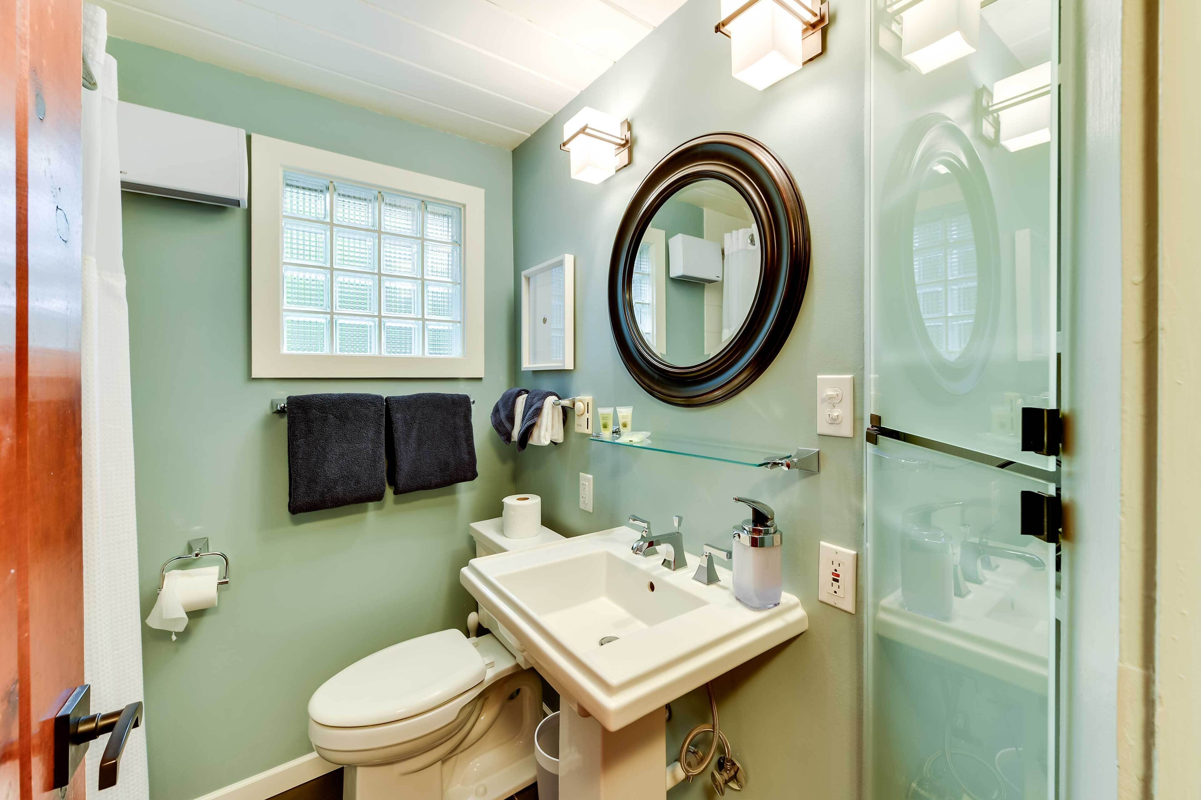 A bathroom with a toilet, sink, and mirror. There is a window with blinds, a towel rack, and a glass shower enclosure.
