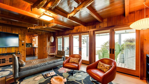 Ein gemütliches Wohnzimmer mit Holzwänden, einem großen Flachbildfernseher, Ledersofas und einem großen Fenster mit Blick auf Pflanzen.
