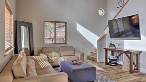 Ett vardagsrum med en beige soffa, en blå pall, ett träbord, en tv som är monterad på väggen och en trappa som leder till ett annat våningsplan.