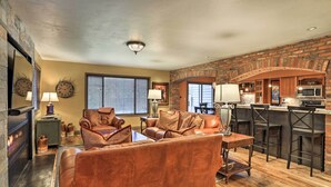 A living room with a fireplace, leather sofa, and a kitchen area with a brick wall and bar stools. - Modern Home w/ Patio: 3 Mi to Beaver Creek Resort! (Avon)