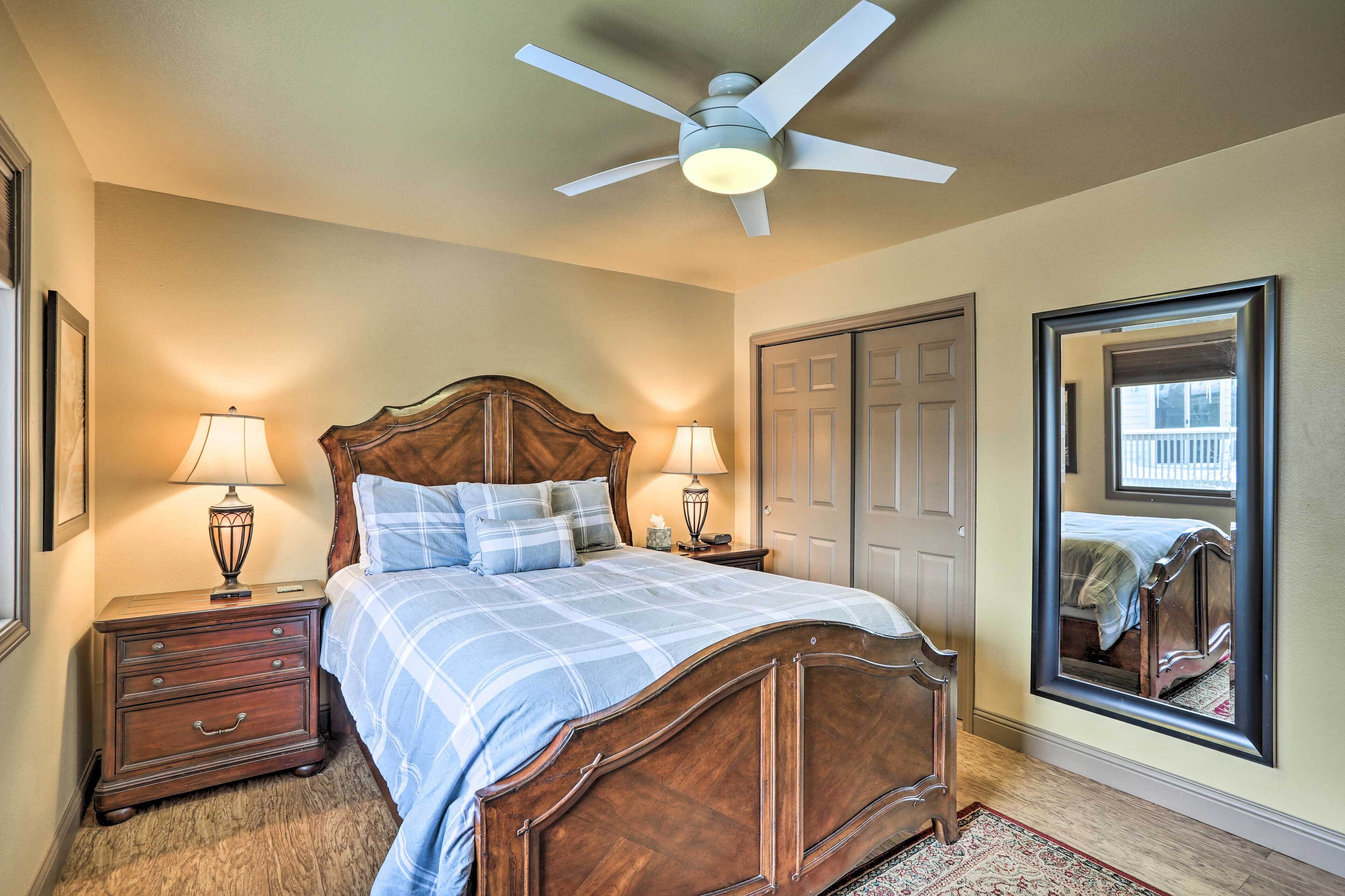 A bedroom with a wooden bed, two bedside tables with lamps, a ceiling fan, a large mirror, and a door.