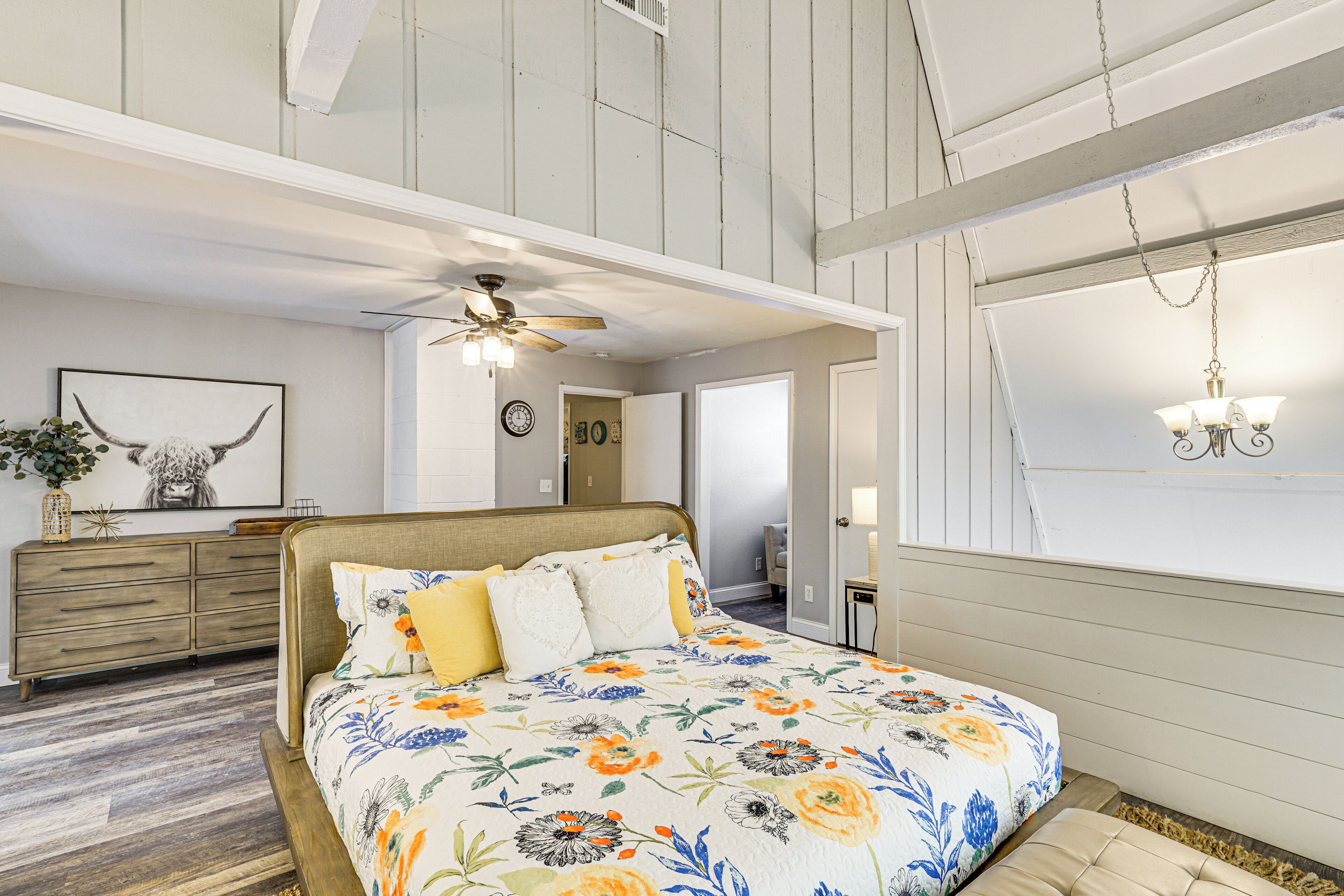 A bedroom with a bed, a ceiling fan, a chandelier, a dresser, and a window with a view.