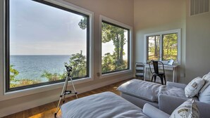 Ein Zimmer mit großen Fenstern, die auf ein Gewässer blicken, einem Stativ und einem Schreibtisch mit Stuhl.