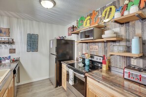 Una cucina con mobili in legno, un frigorifero, un fornello e un forno a microonde. Si vedono scaffali con barattoli e un orologio appeso al muro.