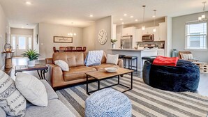 A modern living room with a leather sofa, a coffee table, and a black ottoman. There is a kitchen area with white cabinets and stainless steel appliances. - Spacious Home w/ Patio Near Fishers! (Fortville)