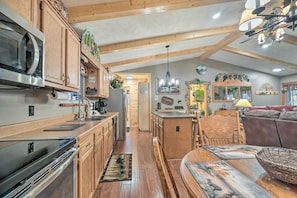 A spacious kitchen with wooden cabinets, a microwave, and a stove. There is a dining area with a wooden table and chairs, and a sofa. The room has wooden beams on the ceiling and various decorations on the walls.