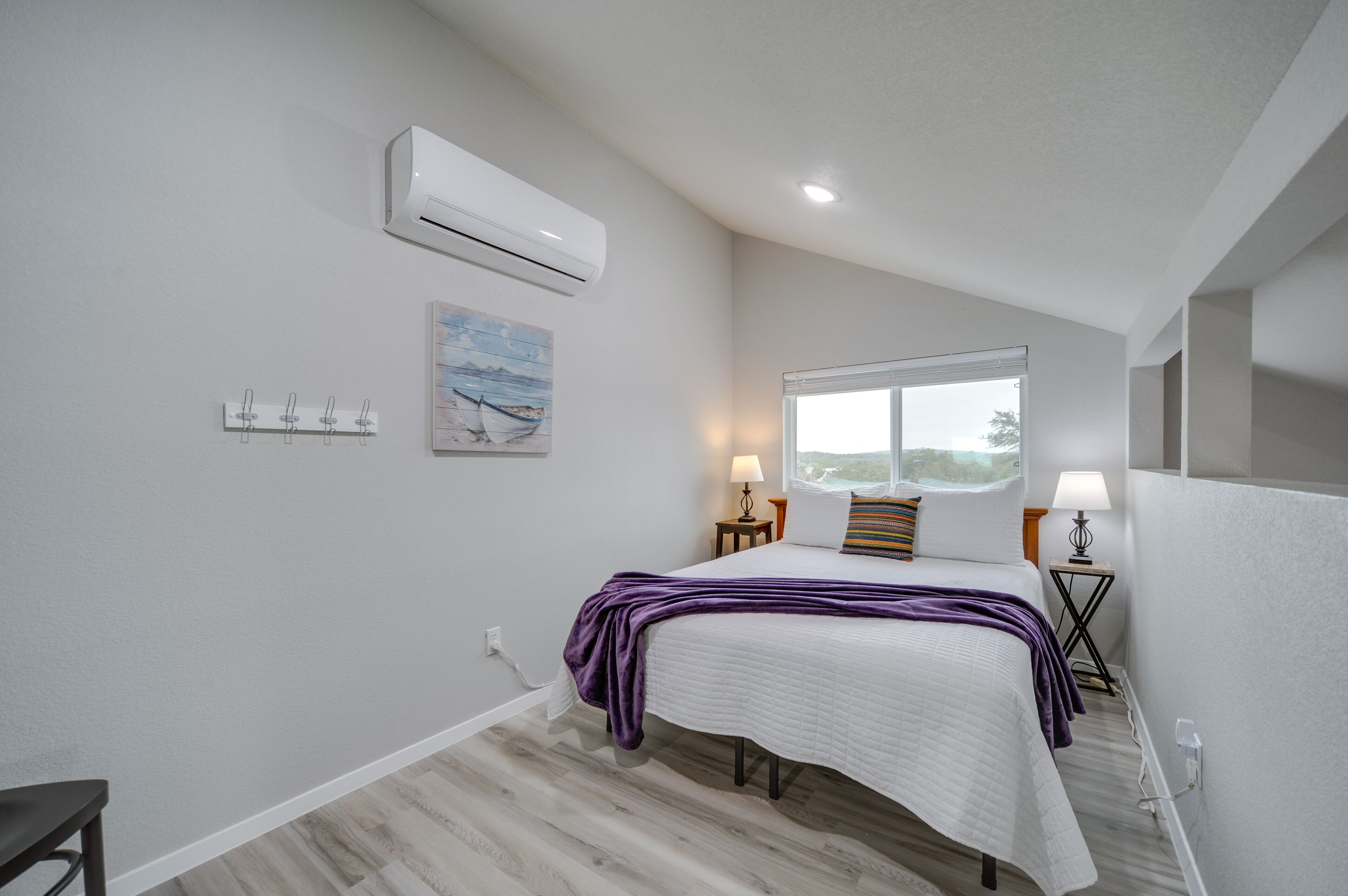 A bedroom with a bed, a window, an air conditioner, a painting, and a lamp.