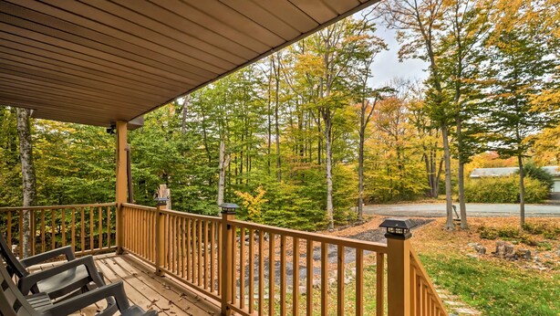 A covered deck with wooden railings and chairs, overlooking a forested area with autumn foliage.