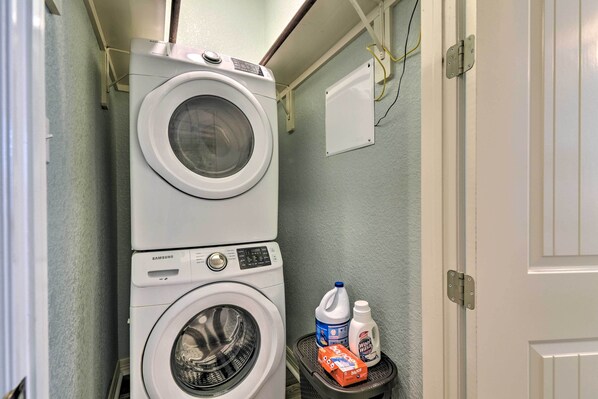 Un espace réduit dédié à la buanderie, comprenant une machine à laver et un sèche-linge superposés, une petite étagère avec des produits de nettoyage et une porte à cadre blanc.