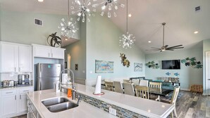 A modern kitchen with white cabinets, a stainless steel refrigerator, and a large island with a sink. There are ceiling fans, artwork on the walls, and a dining area with chairs and a table. - Private Beach + Oceanfront Balcony: Freeport Home (Freeport)