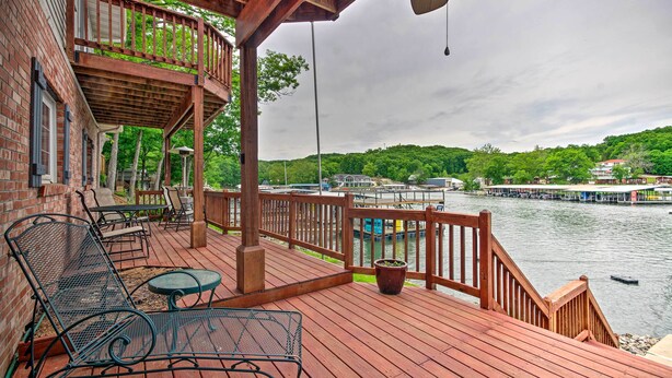 A wooden deck with outdoor furniture, a view of a river with boats, and a forested hillside in the background.