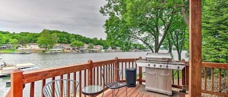 A wooden deck with a grill and chairs overlooking a lake with boats and houses in the background.