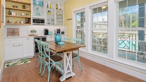 A bright kitchen with white cabinets, a wooden dining table, teal chairs, and a large window overlooking a pool. - Magnolia Cottage by Miss Kitty’s (Rockport)