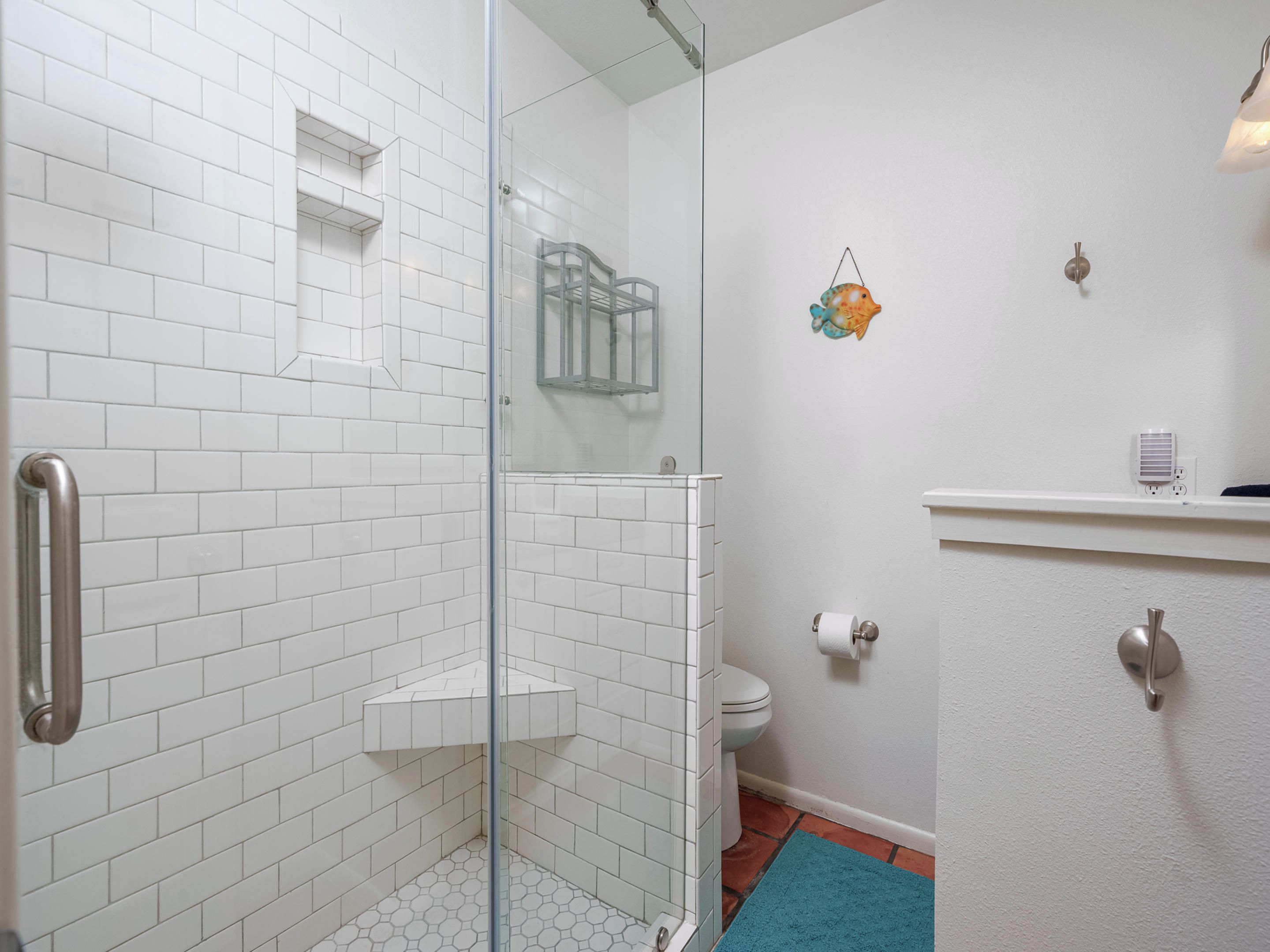 Une salle de bain avec des murs recouverts de carreaux blancs, une douche en verre, des toilettes et un lavabo surmonté d’un tapis bleu.