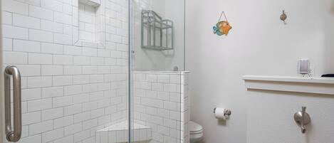 Une salle de bain avec des murs recouverts de carreaux blancs, une douche en verre, des toilettes et un lavabo surmonté d’un tapis bleu.