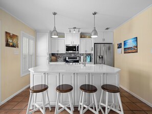 A modern kitchen with white cabinetry, a central island with stools, stainless steel appliances, and pendant lighting. - Pelican Cove by Miss Kitty’s (Rockport)