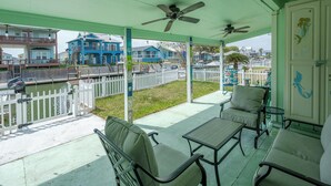 A covered outdoor patio with seating, a ceiling fan, and a view of the water and houses in the background. - Pelican Cove by Miss Kitty’s (Rockport)