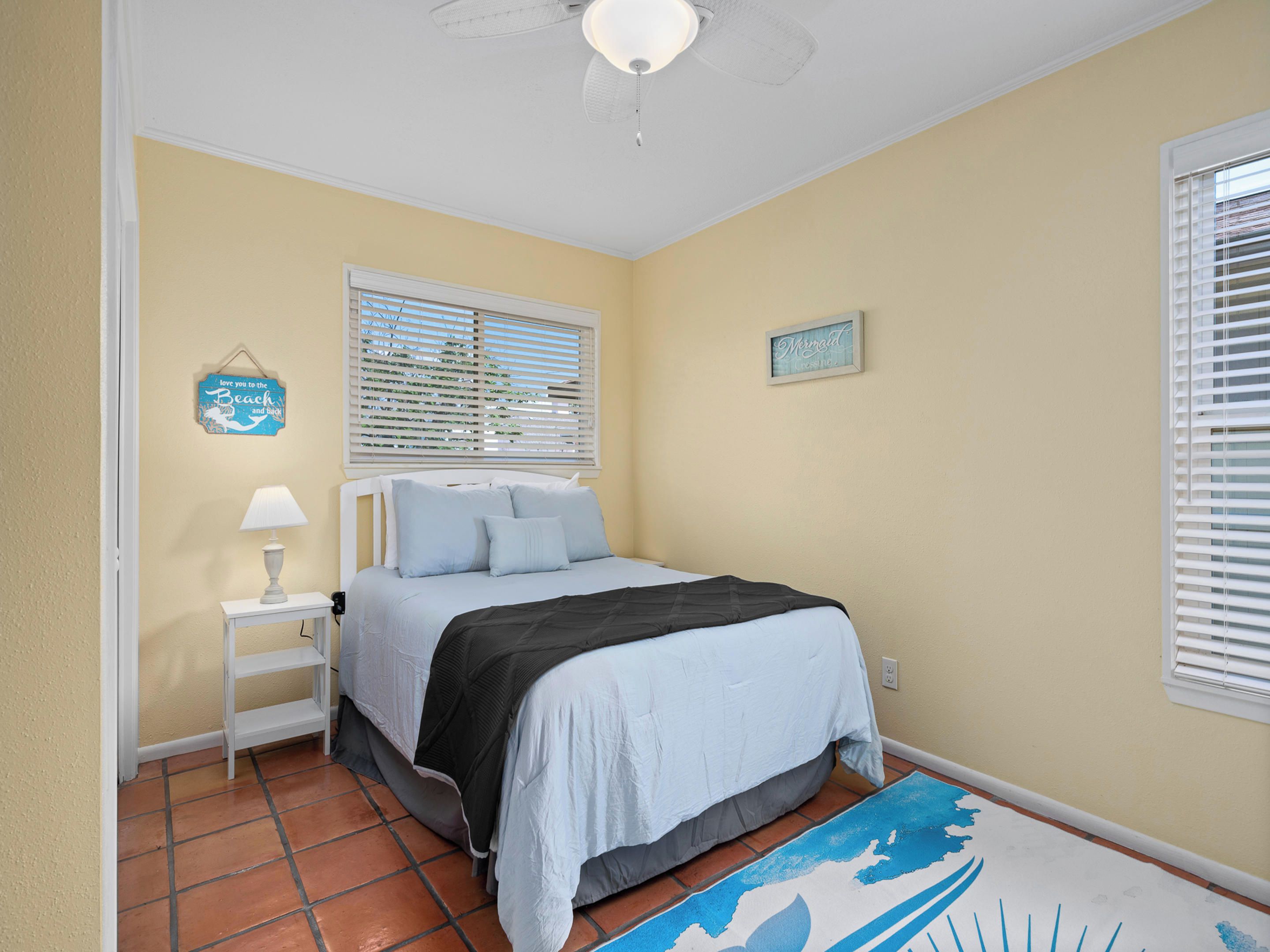 Une chambre à coucher comprenant un lit, une table de chevet, une lampe, une fenêtre avec des stores et un mur orné d’une affiche.