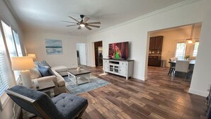 Een ruime woonkamer met een houten vloer, een plafondventilator, een televisie en een keuken op de achtergrond.