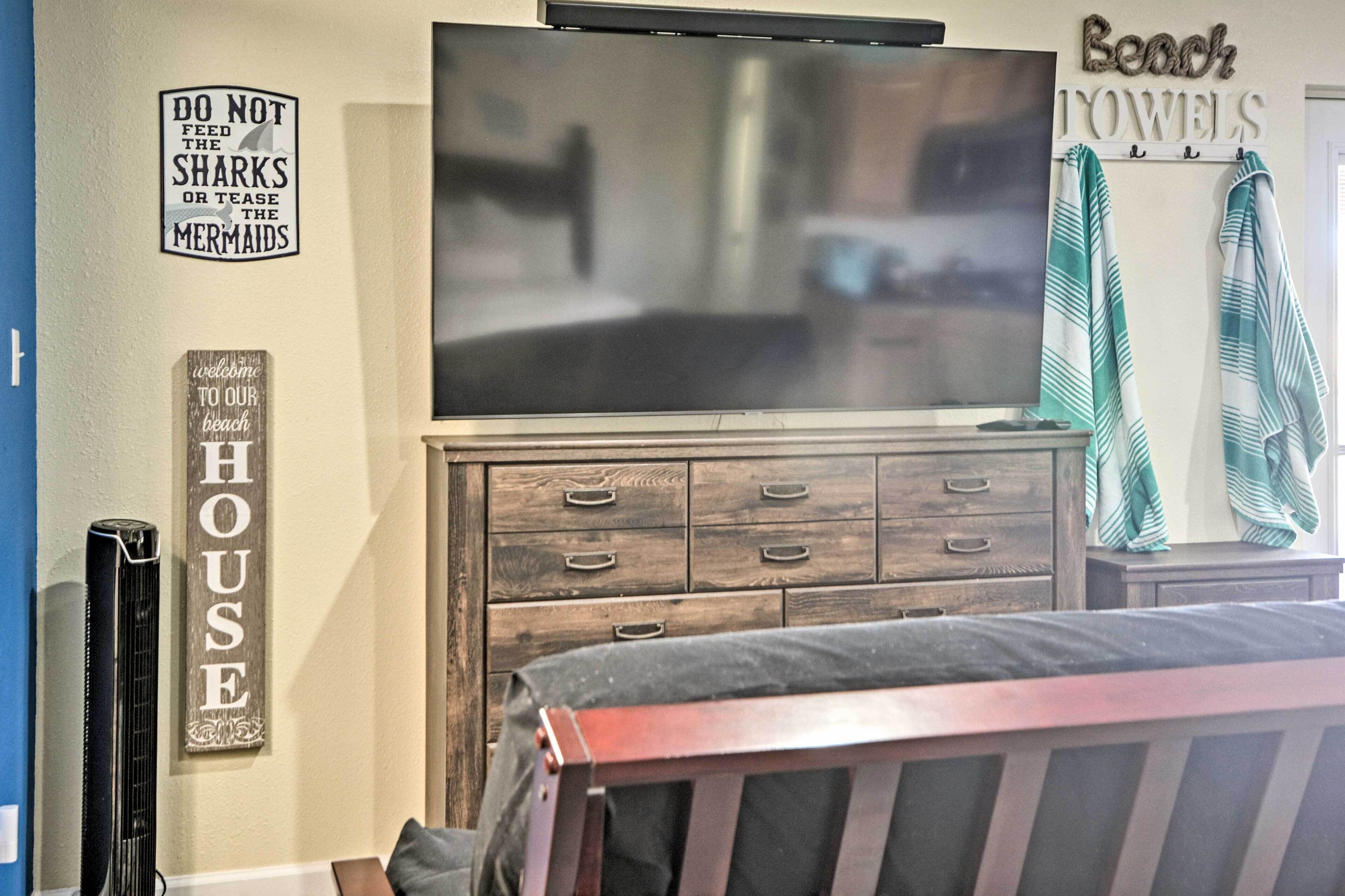 Un dormitorio con una televisión sobre un aparador de madera, una cama con cabecero y adornos en la pared, incluyendo un cartel que dice 'Bienvenido a nuestra casa de playa' y otro cartel que dice 'No alimentes a los tiburones ni molesten a las sirenas'.